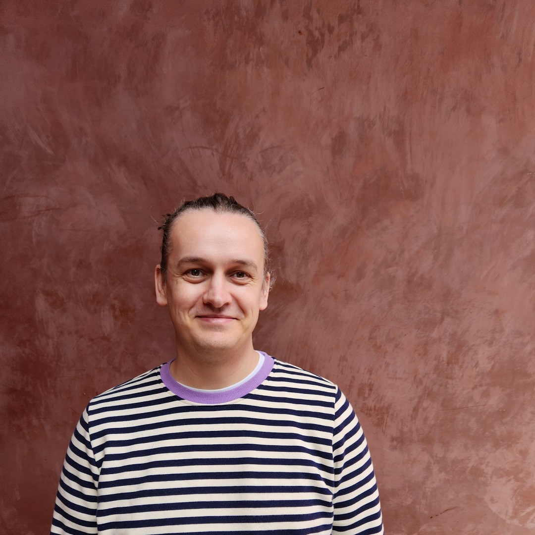 A portrait photo of Ben, they've got their hair up in a bun, are wearing a white and navy striped jumper, and are smiling.